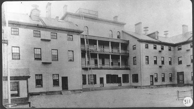 Photo of Tewksbury Almshouse in 1890, a multistory, wooden building with an escape ladder from the 5th story roof to the ground. 