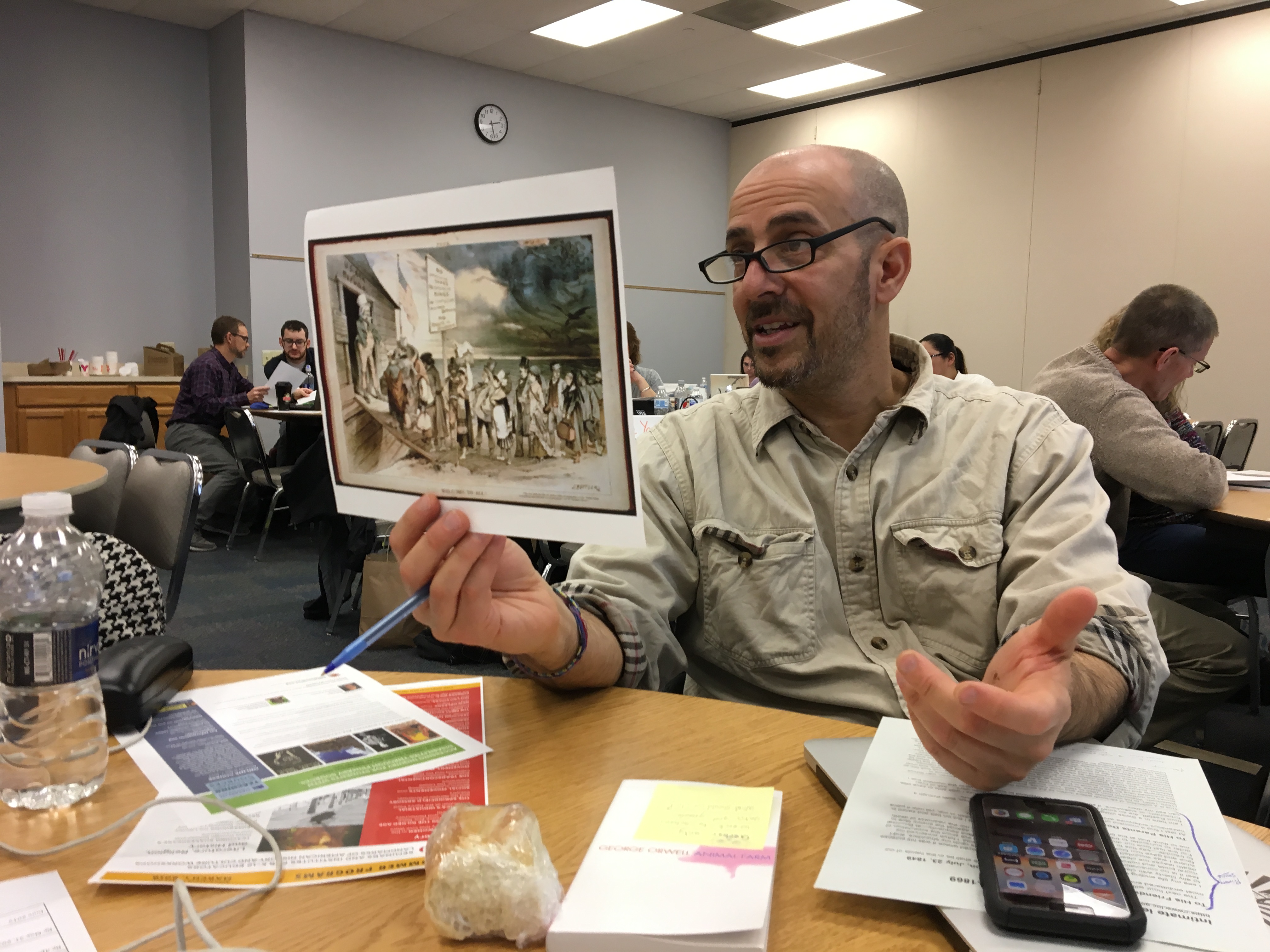Man holds up a color print of an 19th c. cartoon as he gestures with his free hand.