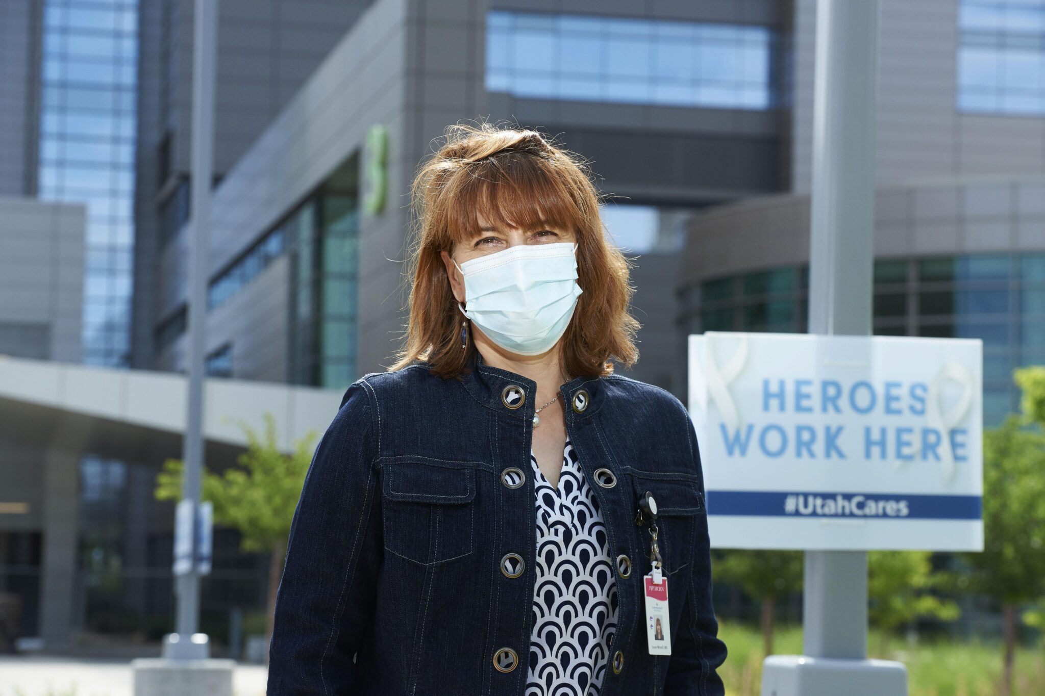 In front of a hospital, a woman in street clothes wearing a medical mask and ID on a lanyard stands in front of a sign saying, "Heroes Work Here."