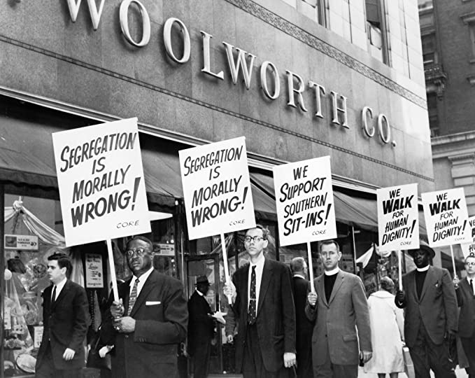 Men in suits, some wearing a minister's clerical collar, carry picket signs reading "Segregation is Morally Wrong" in front of a Woolworths store. 