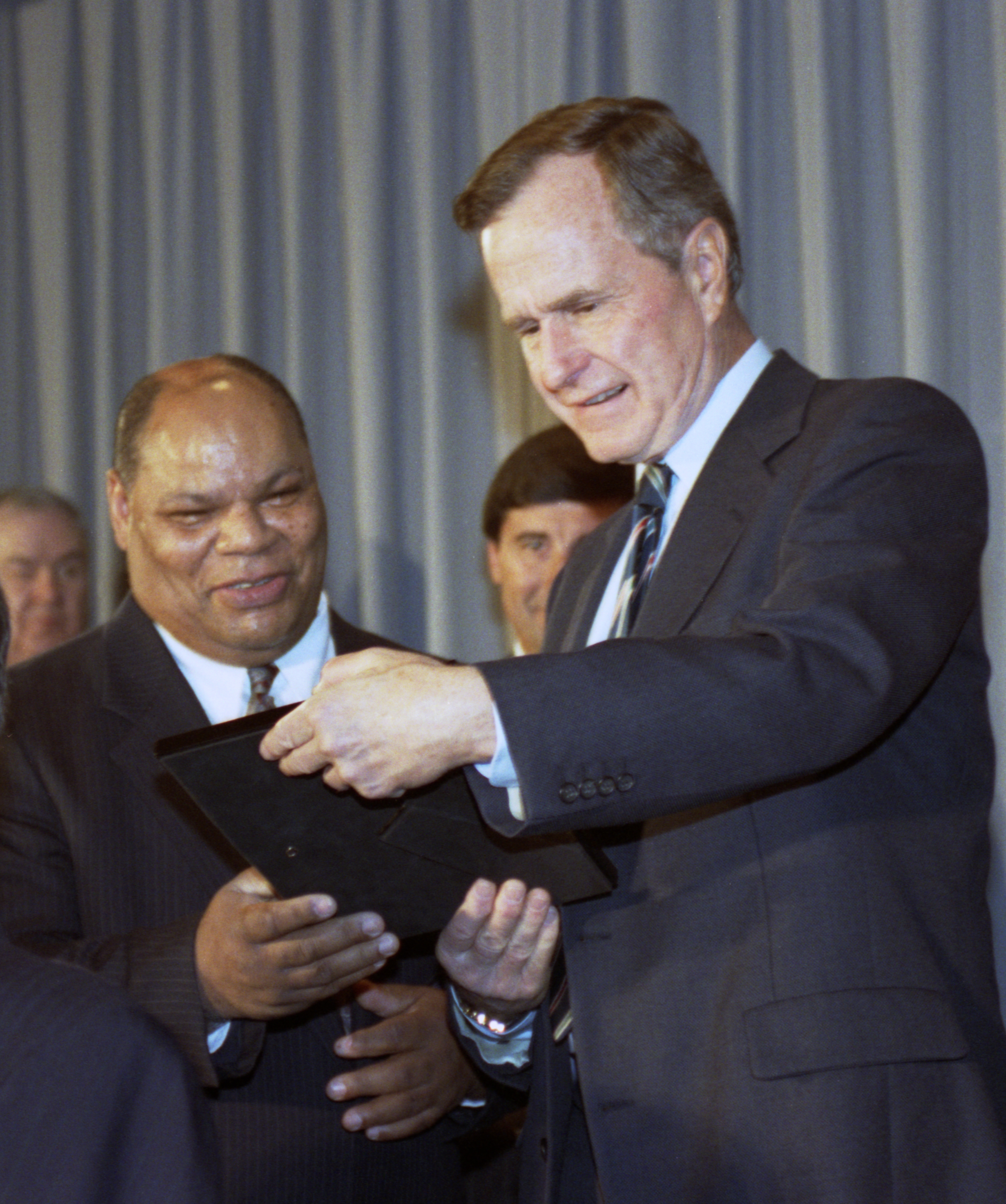 A photograph of President George HW Bush, a tall white man with greying hair wearing a suit, presenting a folder with an ADA award to Ronald Johnson, a shorter African American man with balding hair wearing a suit. The two men stand side by side and look at the award they both have a hand on. 