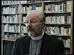 Screen cap from an oral history interview with Jim Munroe. He wears his priests' collar. He is balding and wears glasses. He sits in front of shelves of books. 