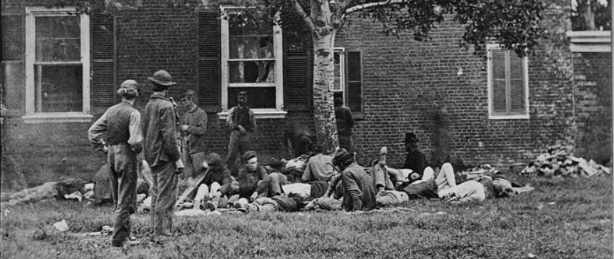 Wounded from the Battle of the Wilderness, some standing, most lying under a tree next to a brick house.