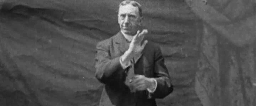 Screen cap from a film of George Veditz wearing a suit and giving a speech in American Sign Language. 