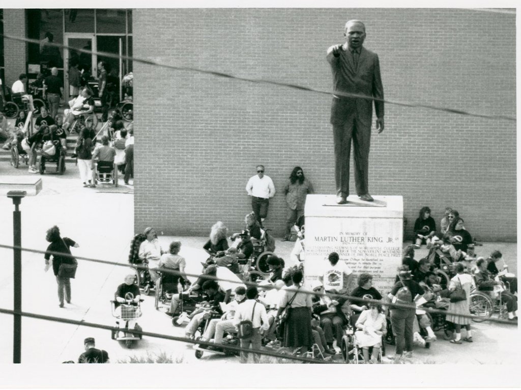 A diverse group of ADAPT activist surround a statue of Martin Luther King, Jr. The activists are young and old, black and white, ambulatory and wheelchair users. The group is gathered around the base of the statue, most of them are sitting around it in a clump. Other activists in the background are at the entrance of a building, showing the lack of accessibility to wheelchair users via the building's steps. 