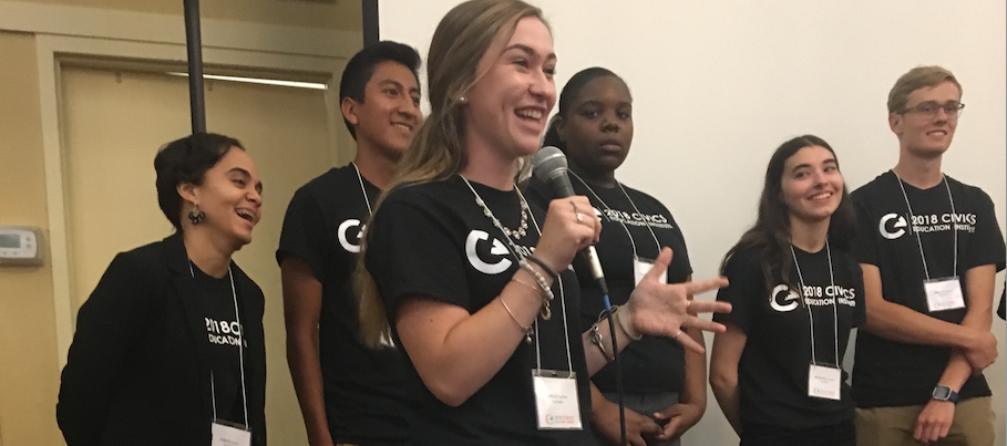 A racially diverse group of high school students smile as they stand in front of the teachers at the 2018 Civics Education Institute. A girl stands in front of the others, speaking into a microphone. 