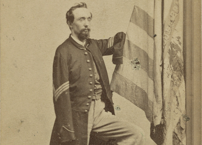 A bearded young man stands in an unbuttoned sergeant's jacket, showing the three chevrons of his rank. Both his sleeves are empty. His right upper arm is raised, resting on the staff of a tattered United States flag.
