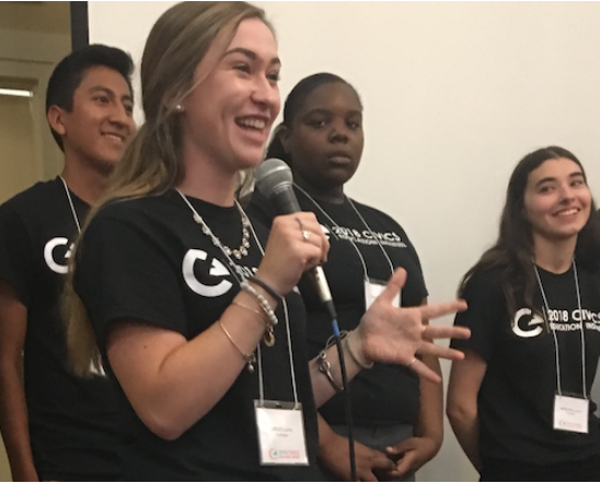 A diverse group of high school students presents on civic engagement at a state educators institute. The speaker has long hair, she is smiling as she holds a microphone. All the students are wearing black civic engagement t-shirts. 