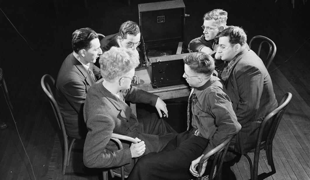 A photo of a group of six men sitting around a table. A large device takes up most of the table space. It is a box that opens up and has a record player inside as well as speakers fo amplify sound. The men sit in chairs and face the device. While some of the man wear glasses, all of their eyes are closed. The men lean towards the device to listen, leaning on their hands and resting their elbows on the table. 