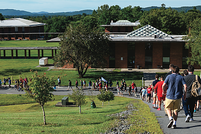 A line of people in informal dress walk across the tree-lined campus of Landmark College. Mountains frame the horizon.
