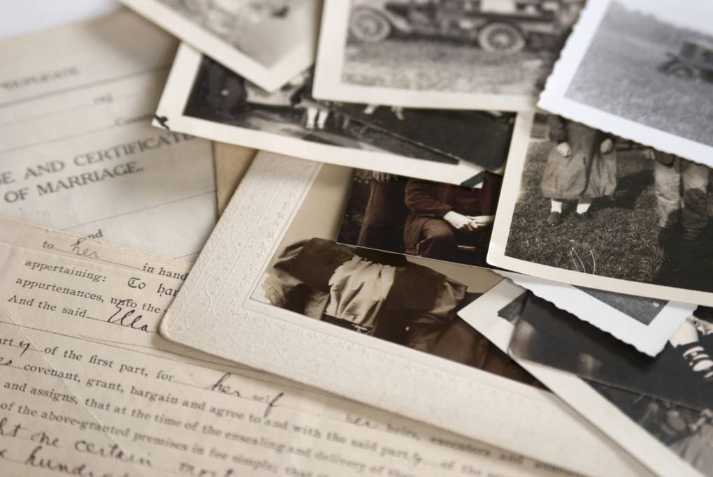 A stack of old photos and forms, including a marriage license, are spread across a table. 