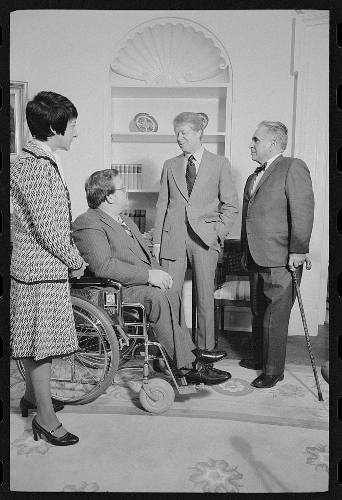 President Carter stands as he talks with a man with a cane, a man in a wheelchair, and a woman. 