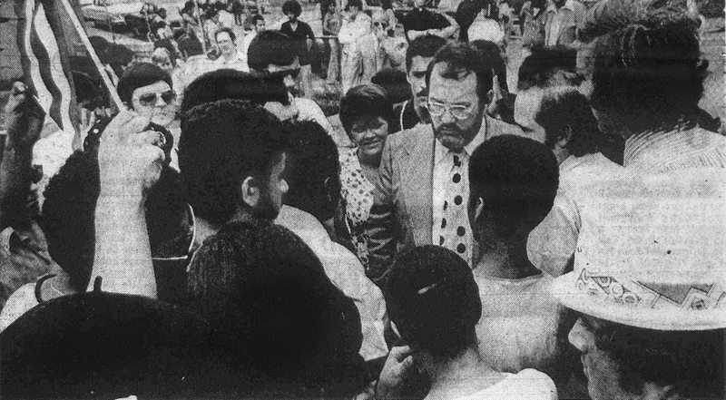 A man in a suit and tie is surrounded by a diverse group of dark-skinned people talking to him.