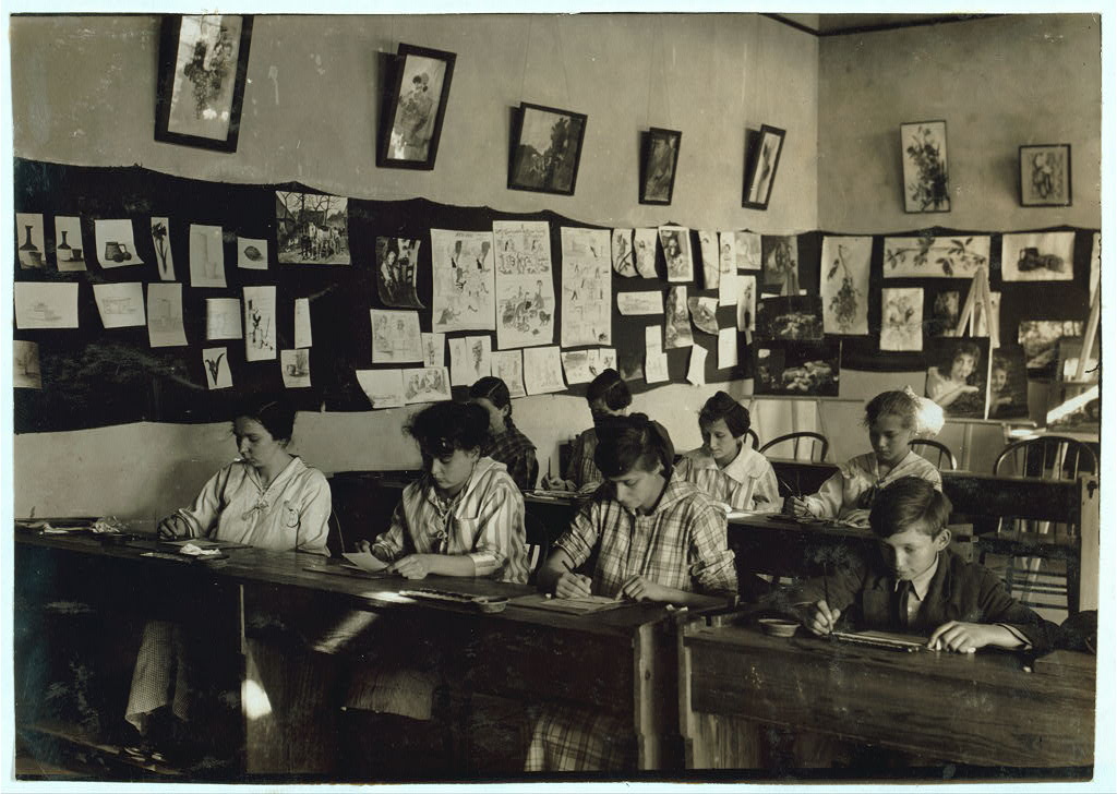 Eight students bend over their desks to draw in 1917 photo. All are well-dressed. The walls are covered with small pictures of artworks.