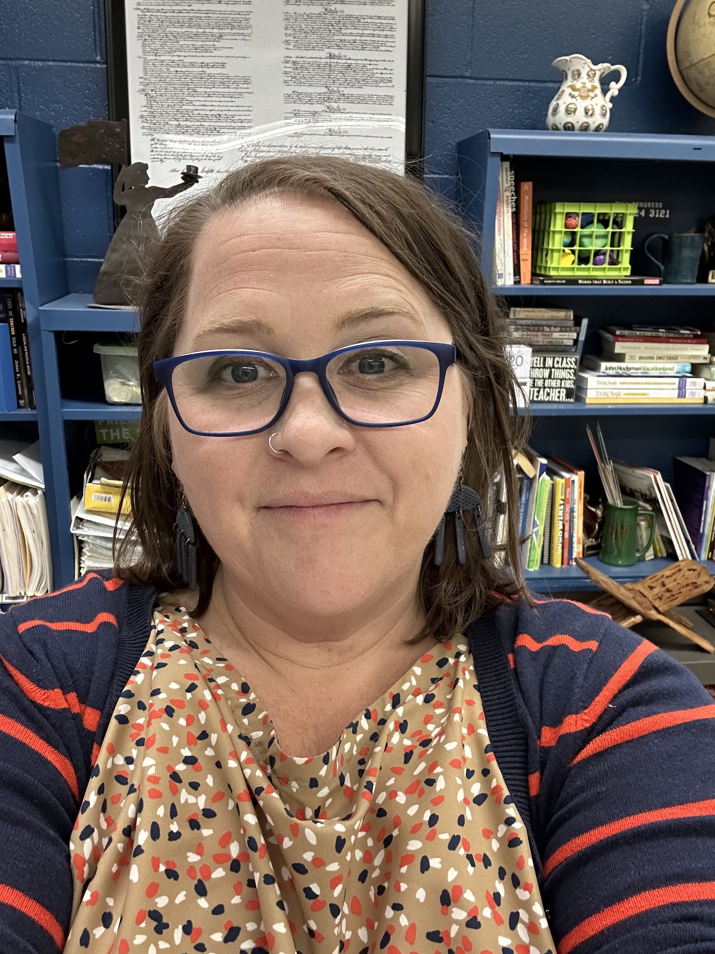 A white woman with shoulder length hair and glasses sits in her classroom in front of a bookshelf with teaching materials and academic books