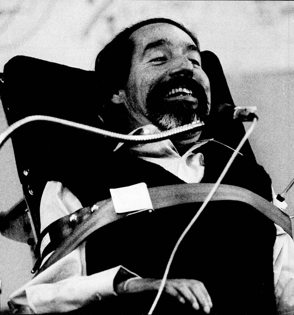 Ed Roberts, a smiling man with dark hair and beard is strapped into his wheelchair. He is speaking into a microphone.
