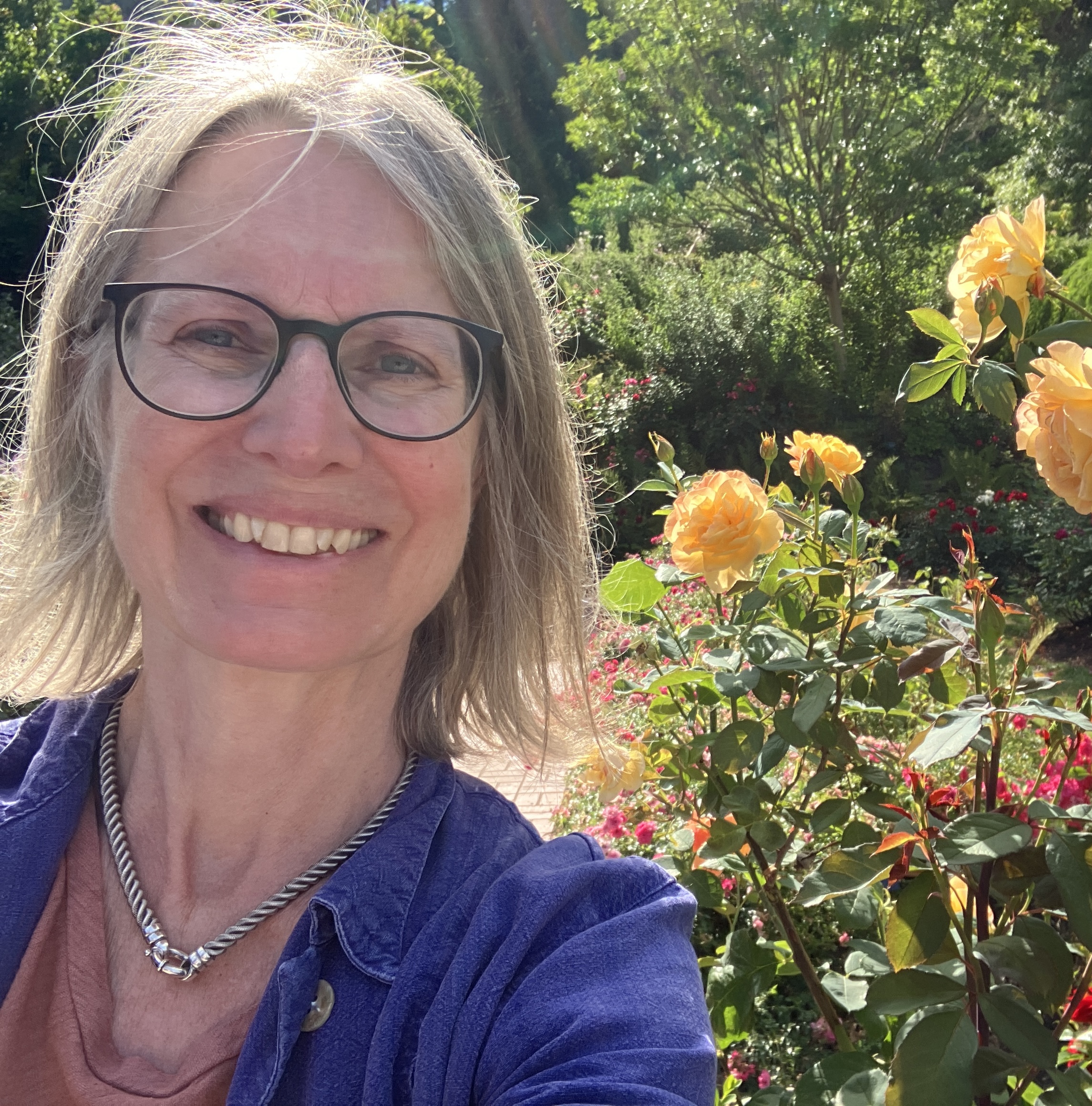 Alison Noyes, a white woman with glasses and light hair, smiles in front of a sunny rose garden.