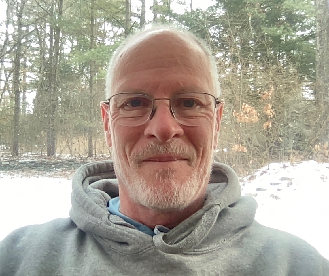 Rich Cairn, a white man with a white beard and glasses, wearing a sweatshirt and seated before a snowy forest scene