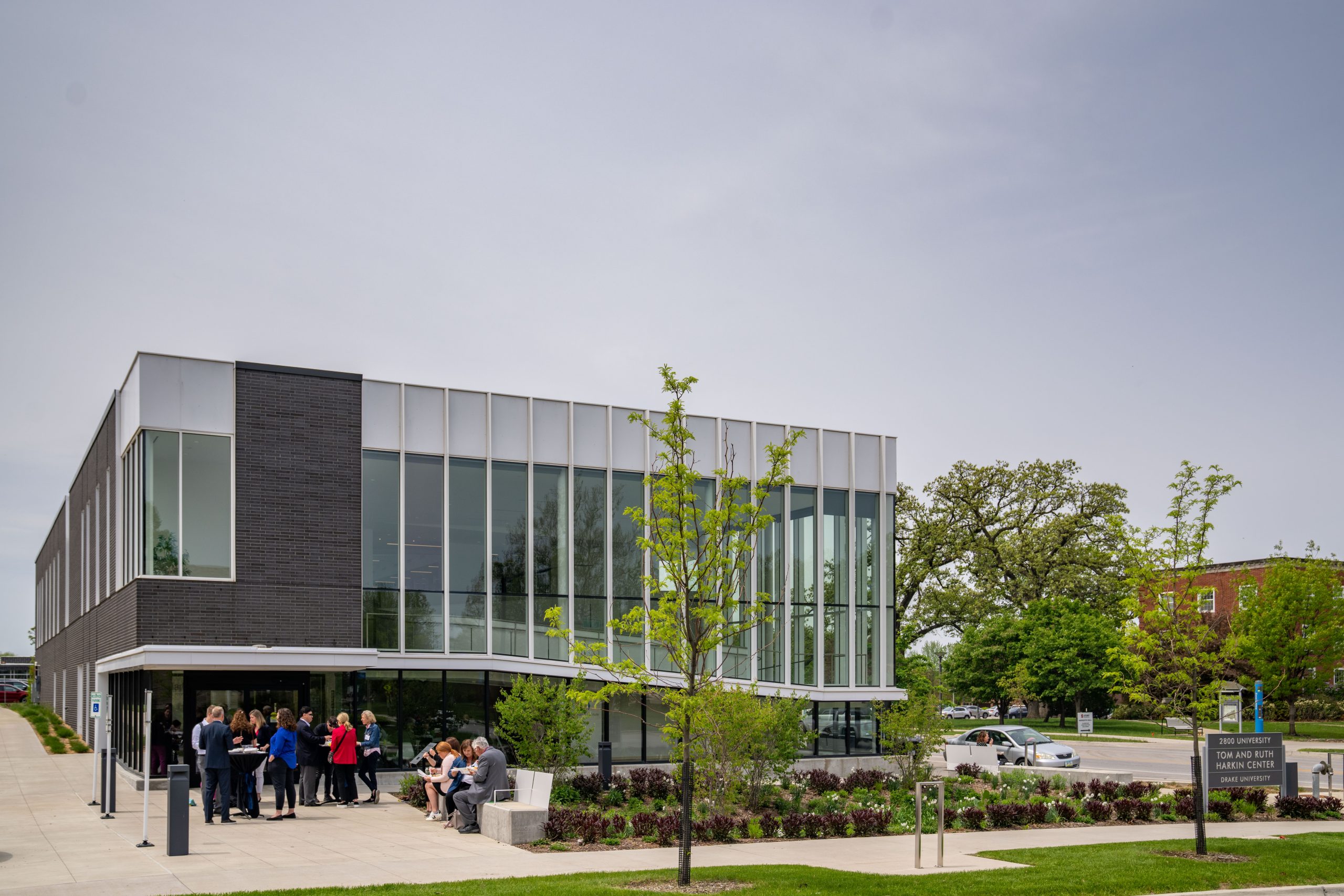 A two story, modern building of glass and brick with leafing trees around it. A dozen men and women in suits and comfortable conference clothes converse near the entrance. 
