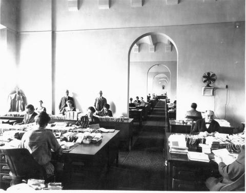 A photo shows ten or so men and women in dresses and suits working at desks with piles of bundled papers. Two men in suits stand and observe. Four more similar offices may be seen through a doorway. 