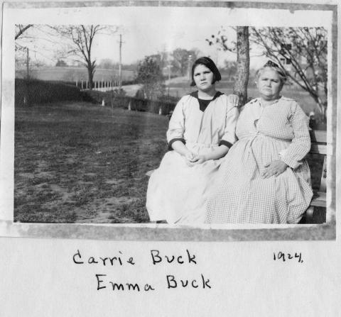 A younger woman in a middy-dress and an older woman in a long dress sit on a park bench on a patchy lawn, a large one-story building visible in the distance behind them. Handwritten in the photo's blank margin: the date 1924 and names Carrie Buck, Emma Buck. 