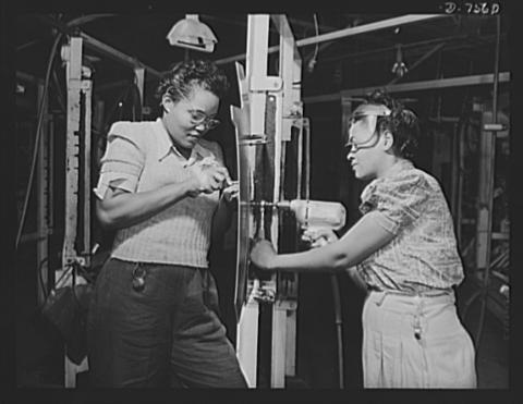 Two black women in street clothes work on an assembly line, one holding a power drill