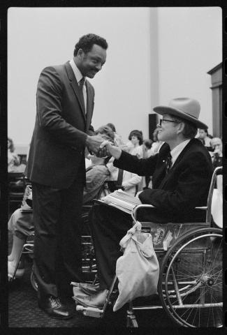 A photo taken in the chambers of the US House of Representatives. Justin Dart, a white man who is sitting in a wheelchair and wearing a suit and a cowboy hat, shakes hands with Reverend Jesse Jackson, an African American man with a mustache who is also wearing a suit.