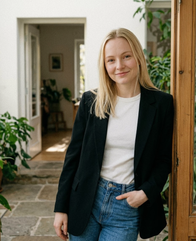 A young woman with long blond hair leans casually against a door frame in the sunroom of a house, with plants and an open door behind her. She wears jeans, a t-shirt and a sports coat.