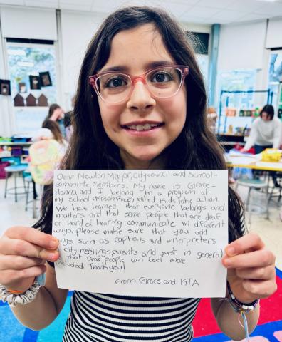 A child with long dark hair and glasses holds a handwritten letter for the viewer to read. She writes to the mayor and other town officials explaining what she has learned about people with disabilities and advocating for interpreters at all town functions.