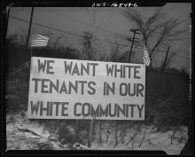 "A large handmade outdoor sign reads "We want white tenants in our white community". The sign is flanked by American flags. "