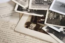 Old photographs and forms, including a marriage certificate are piled on a desk top. 