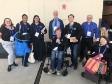 A diverse group of disability activists smile and pose. Five wear Teach Disability History sweatshirts. Two use wheelchairs. All wear conference badges.