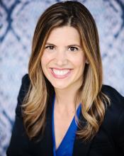 A white woman with shoulder length hair and wearing a blazer smiles at the camera