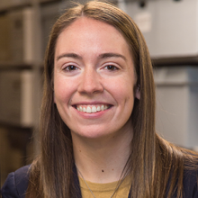 A white woman with straight brown hair wears a suit and smiles.