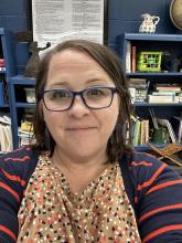 A white woman with glasses sits in a classroom in front of a bookshelf of academic books