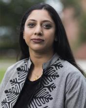 A brown skinned woman with long dark hair wears a loose jacket with a geometric floral pattern.