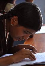 A young Indian woman leans over a desk, eyes closed as she reads a page of braille. She wears long dark hair tied back and has a necklace of beads and a jeweled earring.