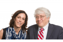 A white woman with dark hair wears a patterned scarf. A white man with white hair and glasses wears a dark suit and striped tie. They sit close to one another and are smiling.