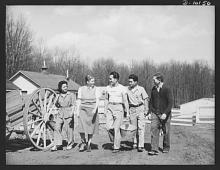 A white woman and man lead a Japanese American woman and two men on a tour of a farm - a wagon wheel is beside them, farm buildings in the background