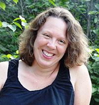 "A smiling woman with wavy brown shoulder-length hair sits before a green hedge. She wears a sleeveless t-shirt.