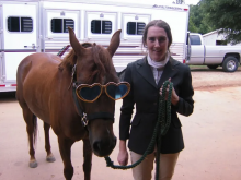 A young white woman in riding clothes holds a rope lead to a horse wearing heart-shaped sunglasses. They stand before a horse trailer.