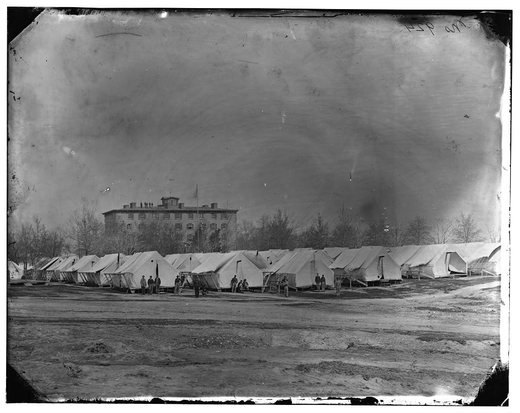 Soldiers lounge in front of dozens of large tents pitched in low rows in front of the Columbia College building. 