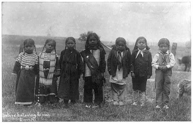 Seven small children, several dressed in Native ceremonial clothes, hair in tight pigtail braids or neatly combed, standing shoulder-to-shoulder in a meadow with a post and wire fencing visible behind them