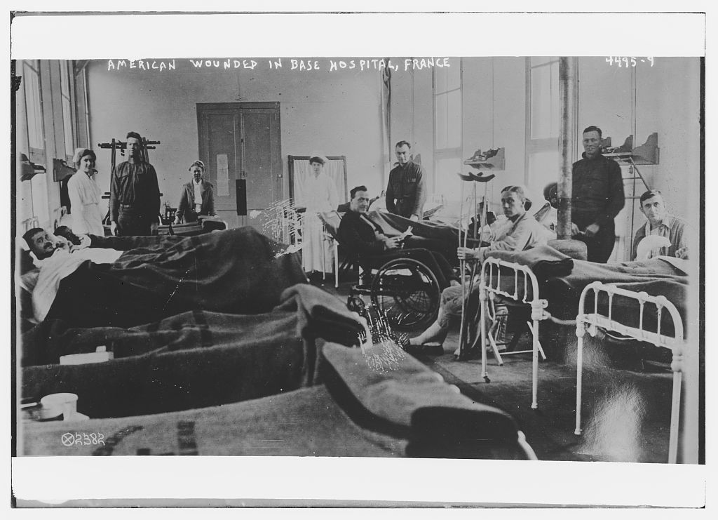A room with at least 5 iron beds with patients lying or sitting up, other convalescing soldiers have crutches, a wheelchair, and a wheeled bed, and 3 white-capped uniformed female nurses and 3 men in soldiers uniforms stand in the background , France,1917 or 1918