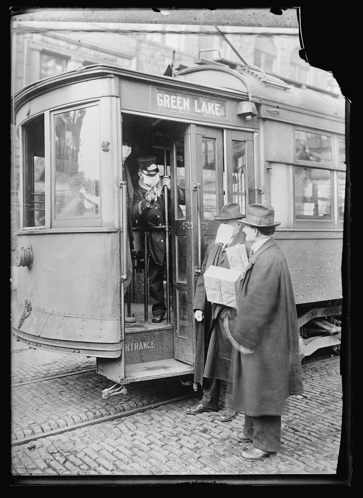 A streetcar with a masked conductor welcoming two passengers in hats and overcoats, one also wearing a mask.