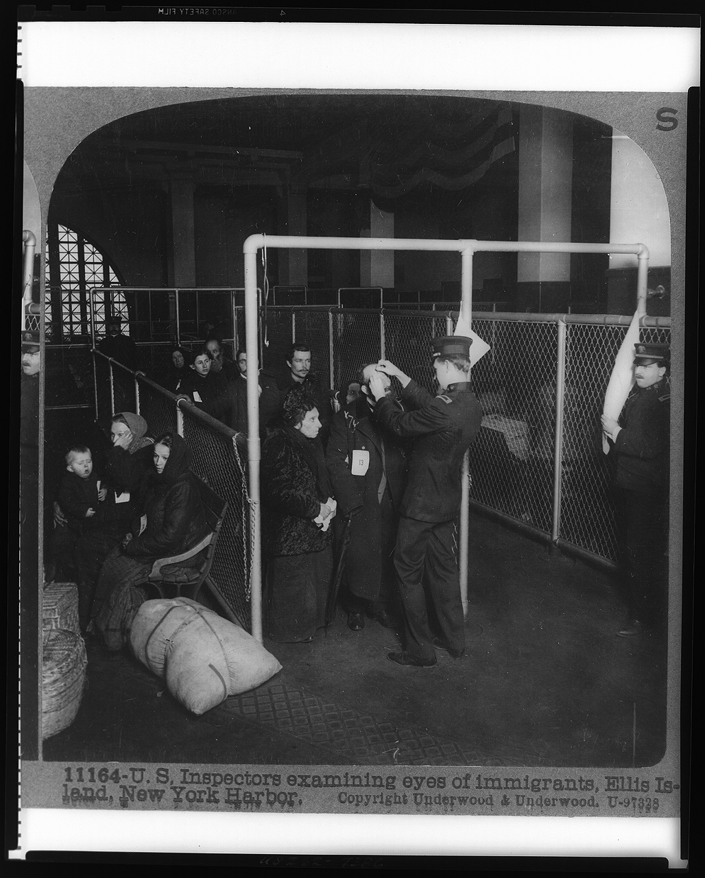 A large hall in Ellis Island Immigration center. Chain linked fence walls divide different lines of immigrants. Two women with a baby sit to the left of the lines on a bench. Two uniformed inspectors stand at the front of the lines, and one uses tools to inspect the eyes of the line of immigrants. Men, women, and children stand in a line with papers pinned to their clothes indicating their immigration information. 
