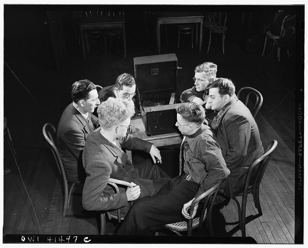 A photo of a group of six men sitting around a table. A large device takes up most of the table space. It is a box that opens up and has a record player inside as well as speakers fo amplify sound. The men sit in chairs and face the device. While some of the man wear glasses, all of their eyes are closed. The men lean towards the device to listen, leaning on their hands and resting their elbows on the table. 