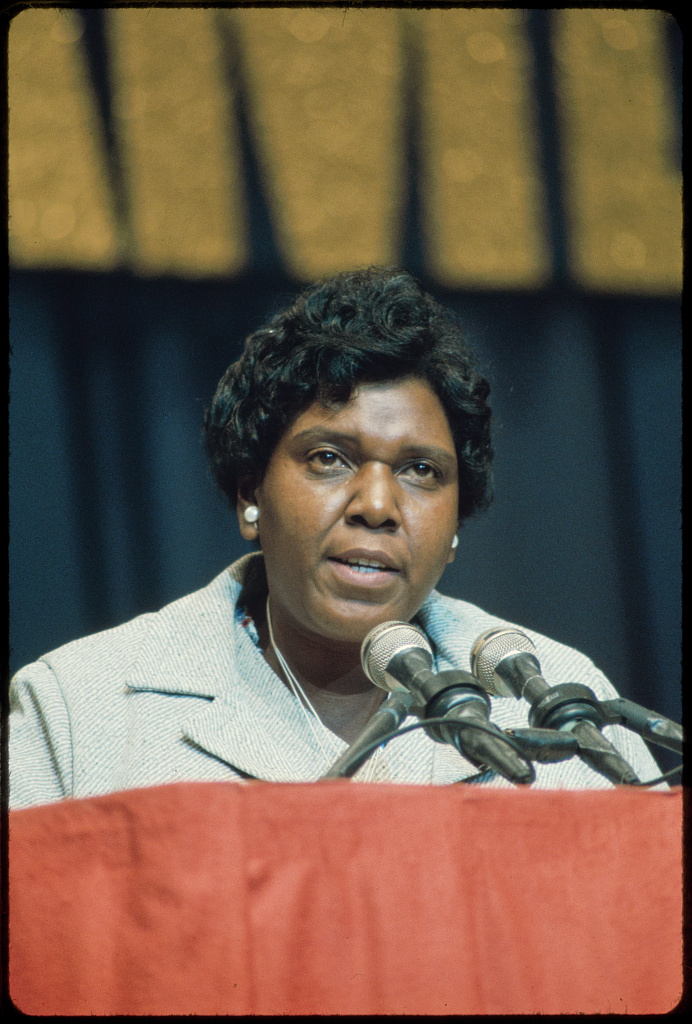 A photograph of Congresswoman Barbara Jordan at a podium speaking into microphones. Congresswoman Jordan is a middle aged African American woman who is wearing a gray coat, large pearl earrings, and has short, wavy, black hair. 