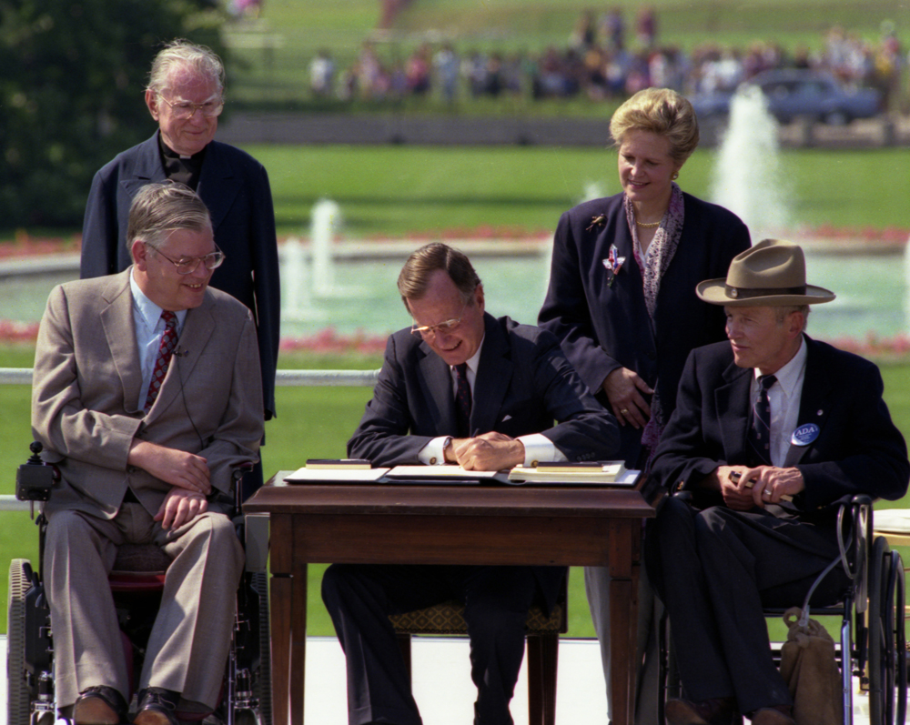 A photograph of President George H.W. Bush signing the Americans with Disabilities Act into law. He is seated at a table outside, there is a fountain in the background. Four other people surround the table, three men and one woman, two men are using wheelchairs. Justin Dart sits to the president's left, wearing his trademark cowboy hat, a suit, and a button that supports the ADA on his lapel. 