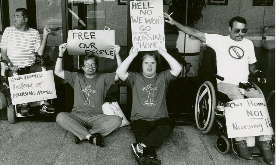 A group of ADAPT activists sitting in wheelchairs or on the ground in front of a building to protest using nursing homes for disabled people. Two wear shirts that say "people first" and they all carry signs that say things like "free our people" or "our home NOT nursing homes."